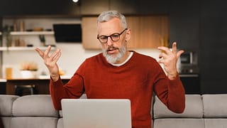 older man at laptop raising his arms as if frustrated