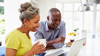 mature man and woman sitting in kitchen looking at laptop computer and smiling couple poc