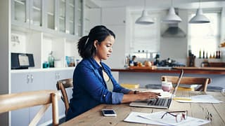 woman at kitchen table with laptop and paperw.width nlFF