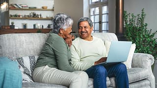two older people sitting on a couch using a laptop and smiling