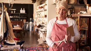 senior woman wearing apron standing next to shelves of apparel in a store small business owner entrepreneur.jpg
