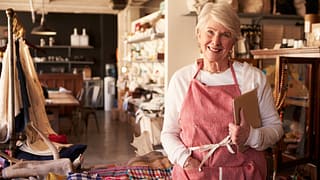 senior woman wearing apron standing next to shelves of apparel in a store small business owner entrepreneur.jpg